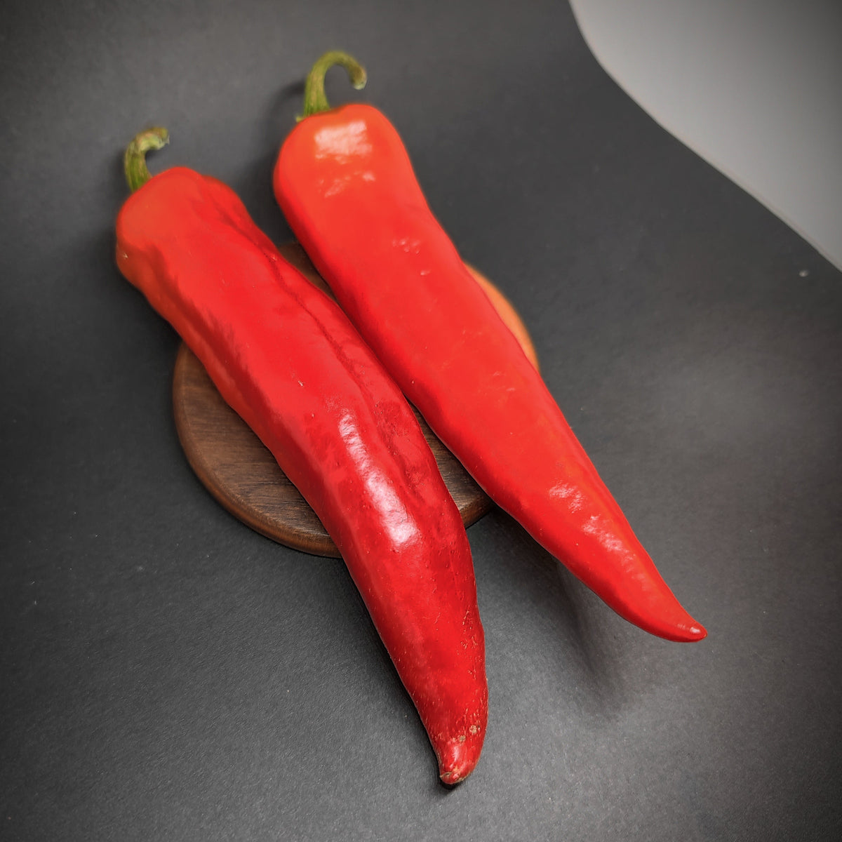 Two long, red Hatch chiles from PepperMerchant.net resting on a small circular wooden base.