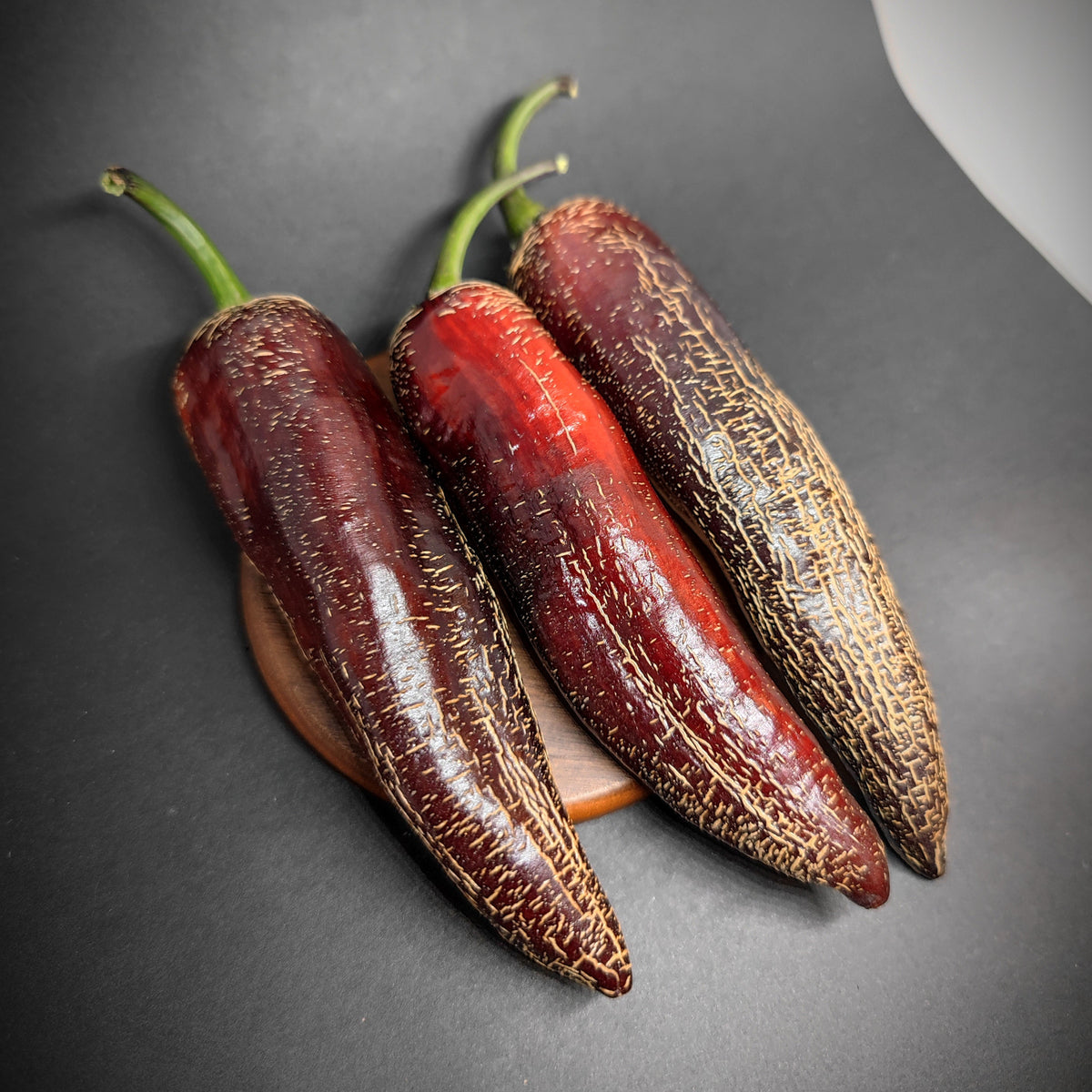Three long, red Dream Catcher x Farmer’s Market Jalapeno peppers with prominent surface corking arranged on a small wooden base.