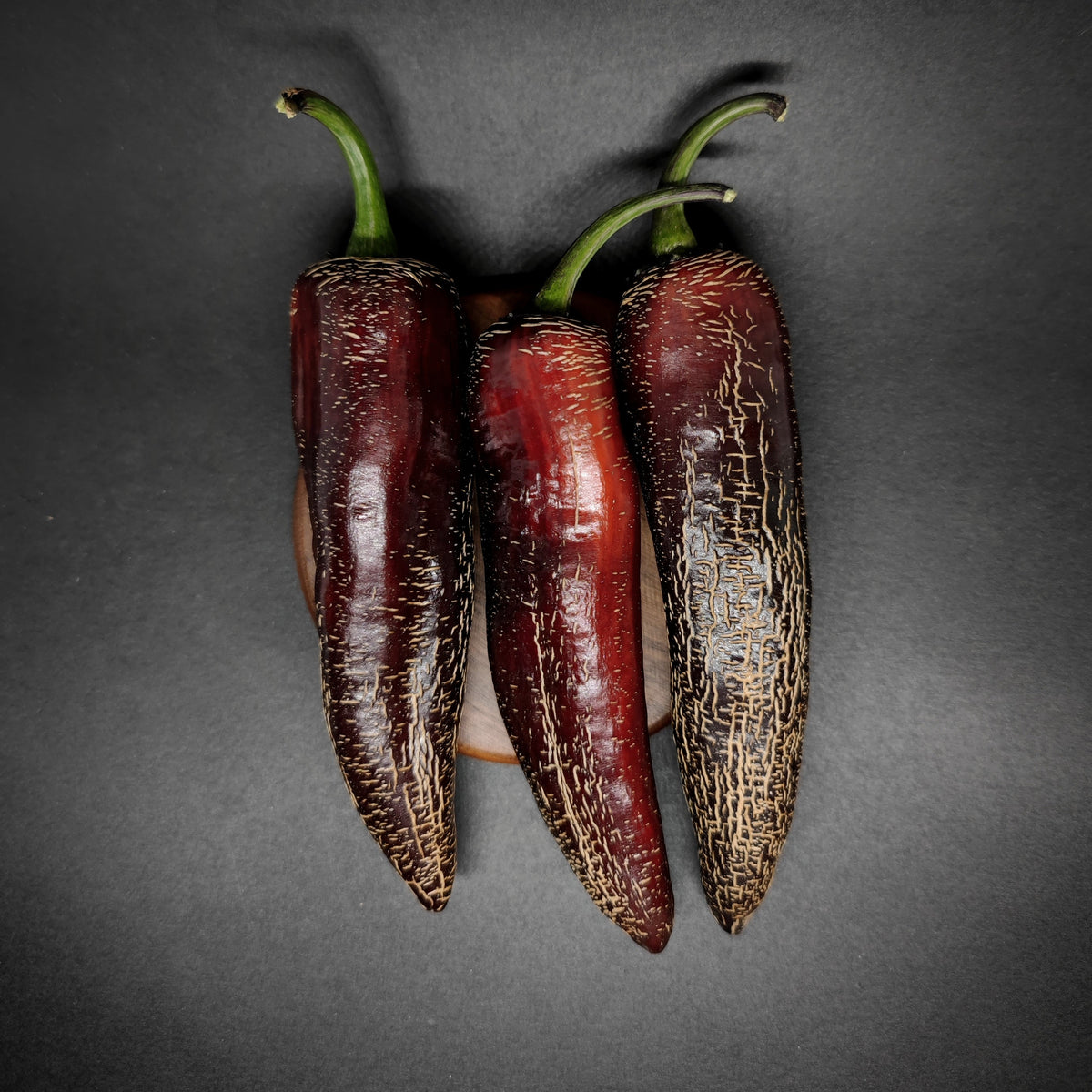 Three dark red, corked Dream Catcher x Farmer’s Market Jalapeno peppers with green stems arranged on a circular wooden base.