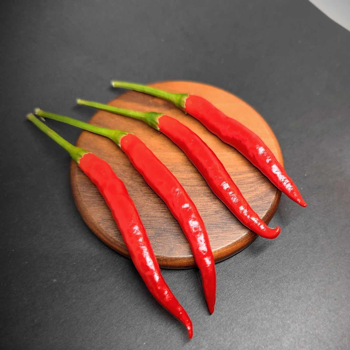 Four red Yatsufusa Japanese chili peppers with green stems arranged diagonally on a round wooden coaster.