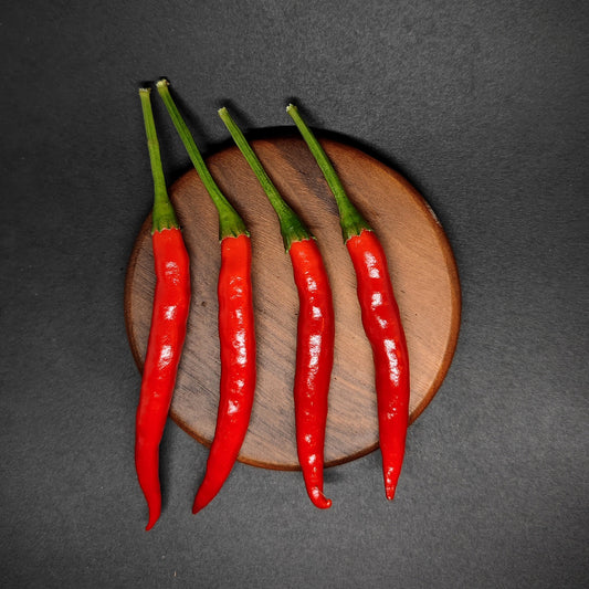 Four bright red Yatsufusa chili peppers with green stems, arranged vertically on a round wooden board.