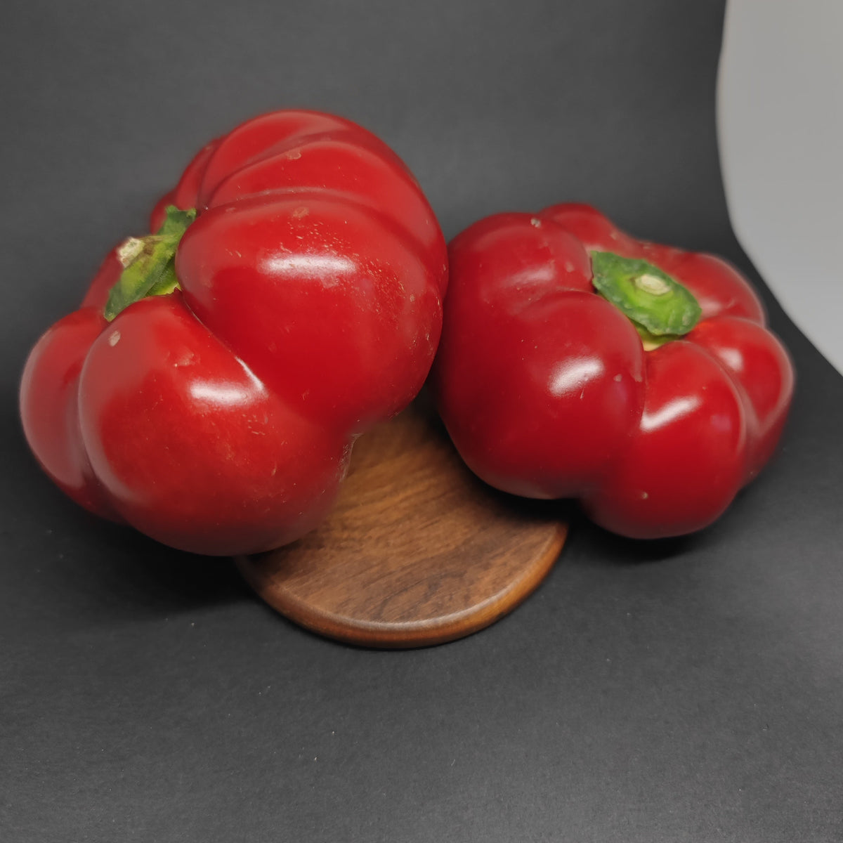 Two bright red, round, lobed Alma Paprika peppers sitting on a small wooden disc.
