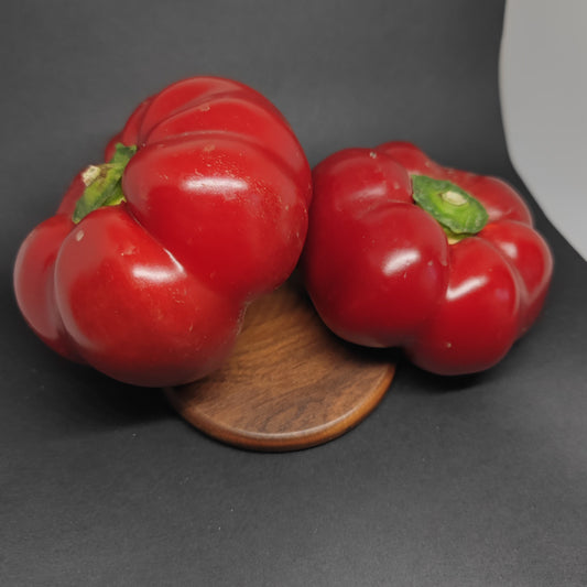 Two bright red, round, lobed Alma Paprika peppers sitting on a small wooden disc.
