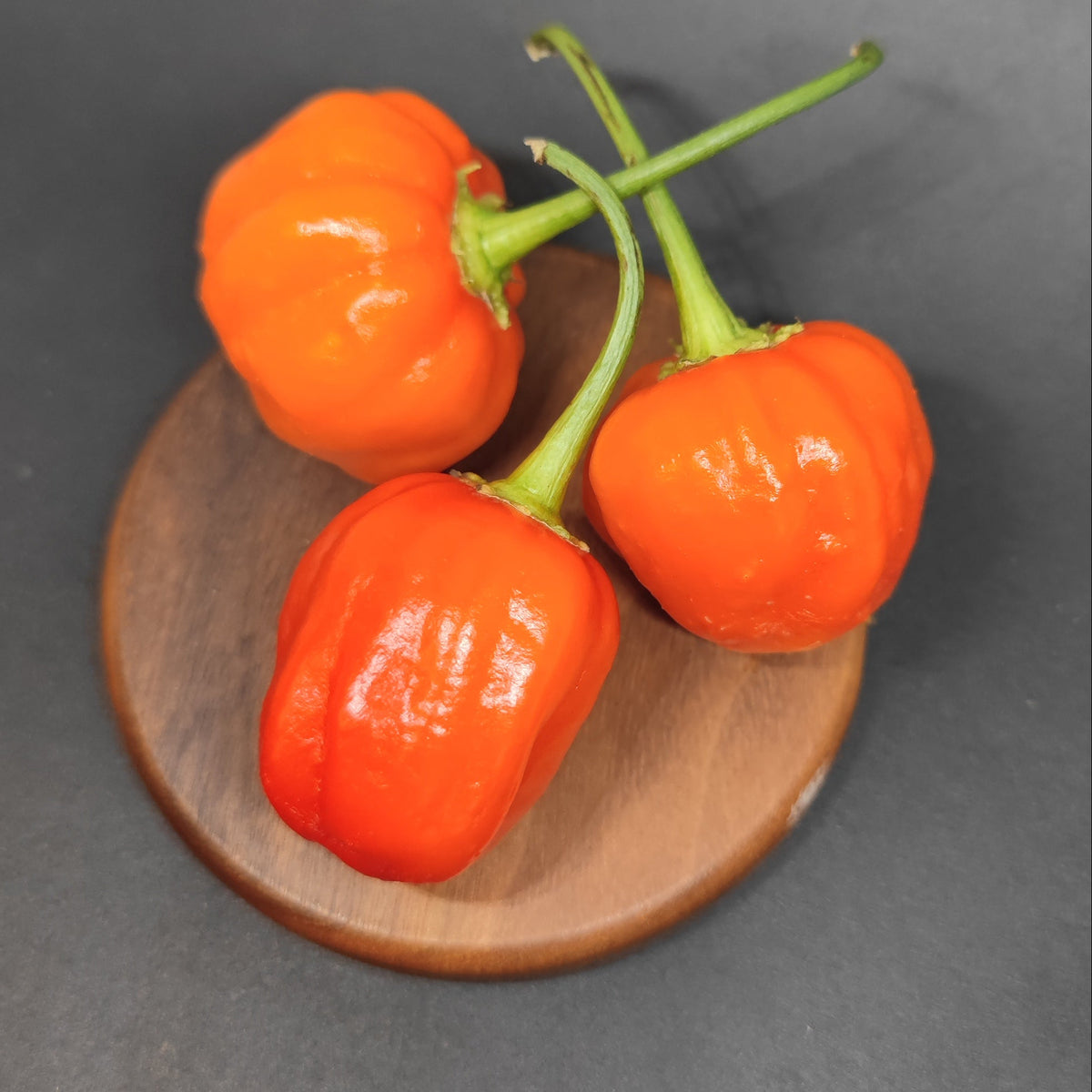 Three bright orange Peppa Peach peppers with green stems rest on a small wooden board.