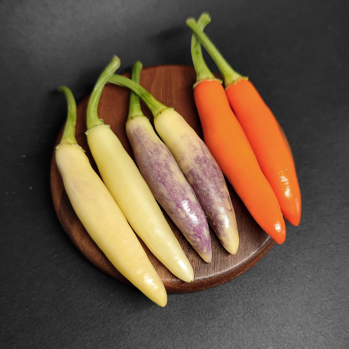 Five slender KS White Thai peppers in shades of cream, purple-streaked white, and bright orange, arranged on a small wooden coaster.