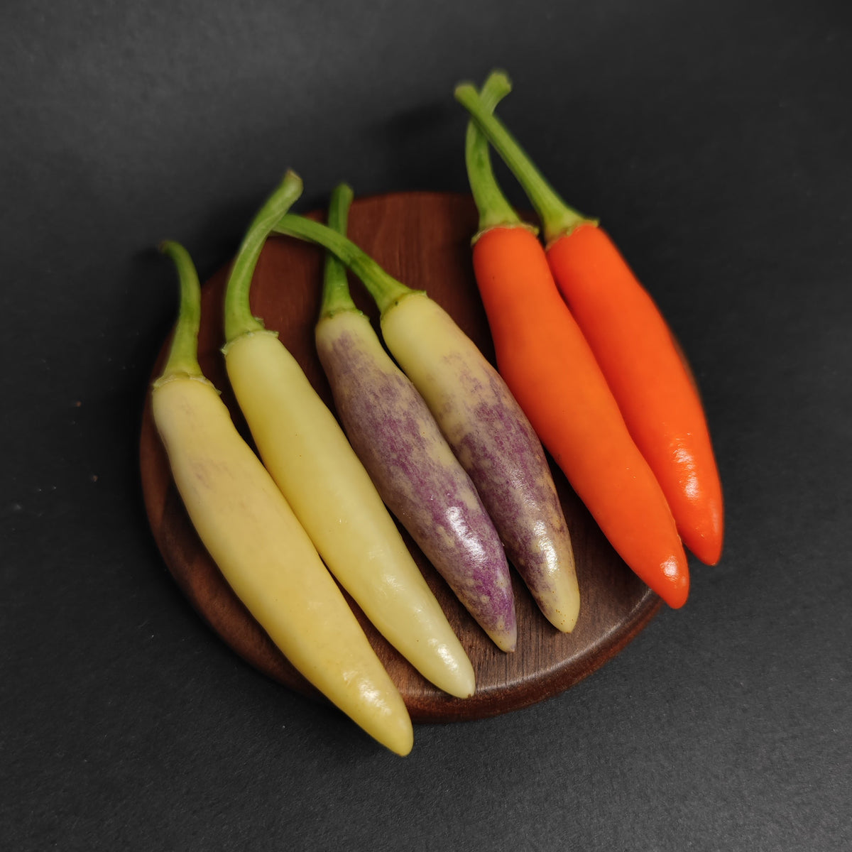 Six KS White Thai peppers ranging from pale yellow to purple-streaked to bright orange arranged on a small wooden coaster.