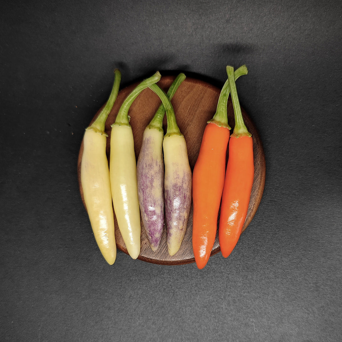 Six chili peppers arranged on a wooden coaster, including KS White Thai, purple-streaked, and orange varieties.