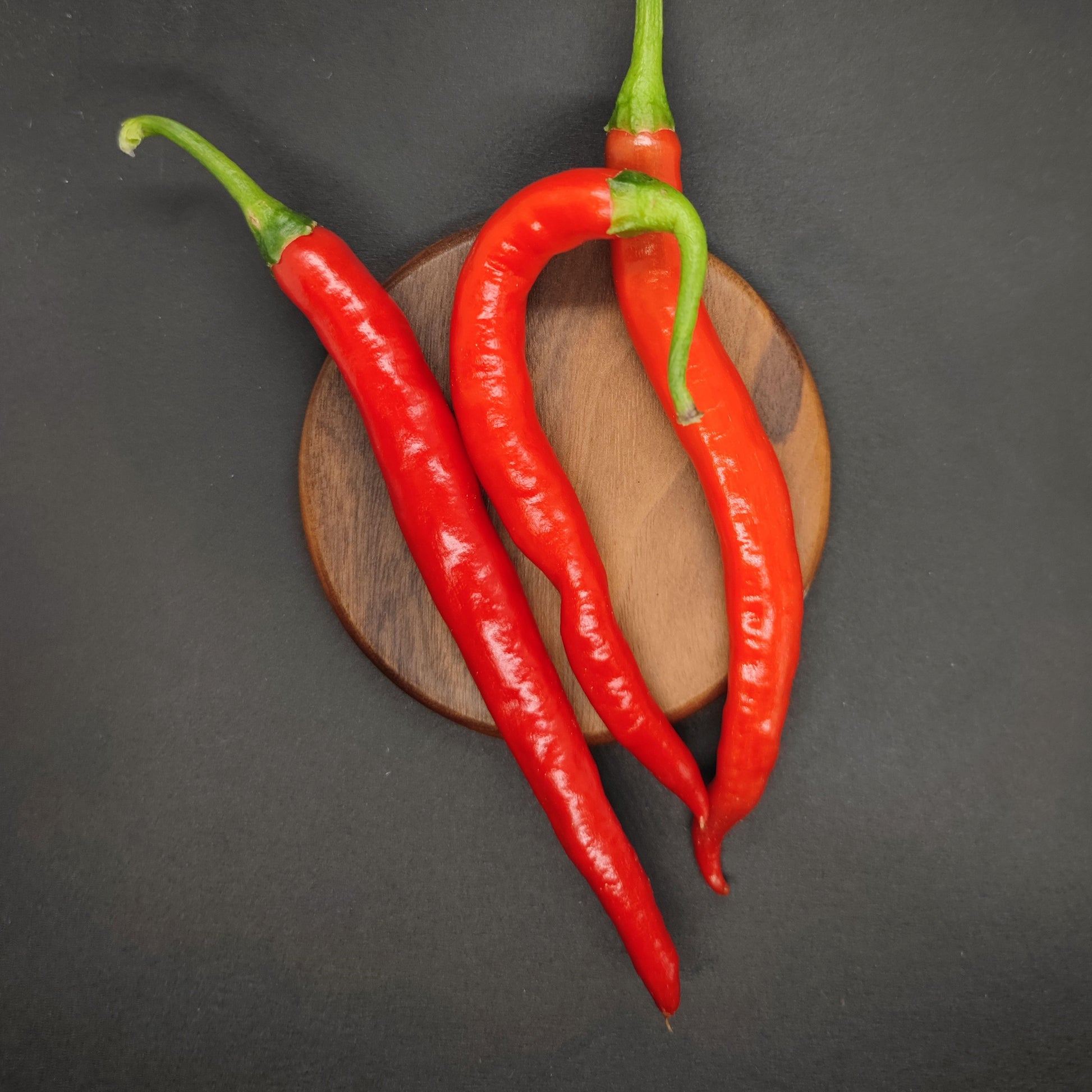 Three vibrant red Hangjiao 10 Afterglow (HJ10) peppers from PepperMerchant.net are arranged on a round wooden board with a black background. The peppers are fresh, long, and slightly curved, with green stems attached.