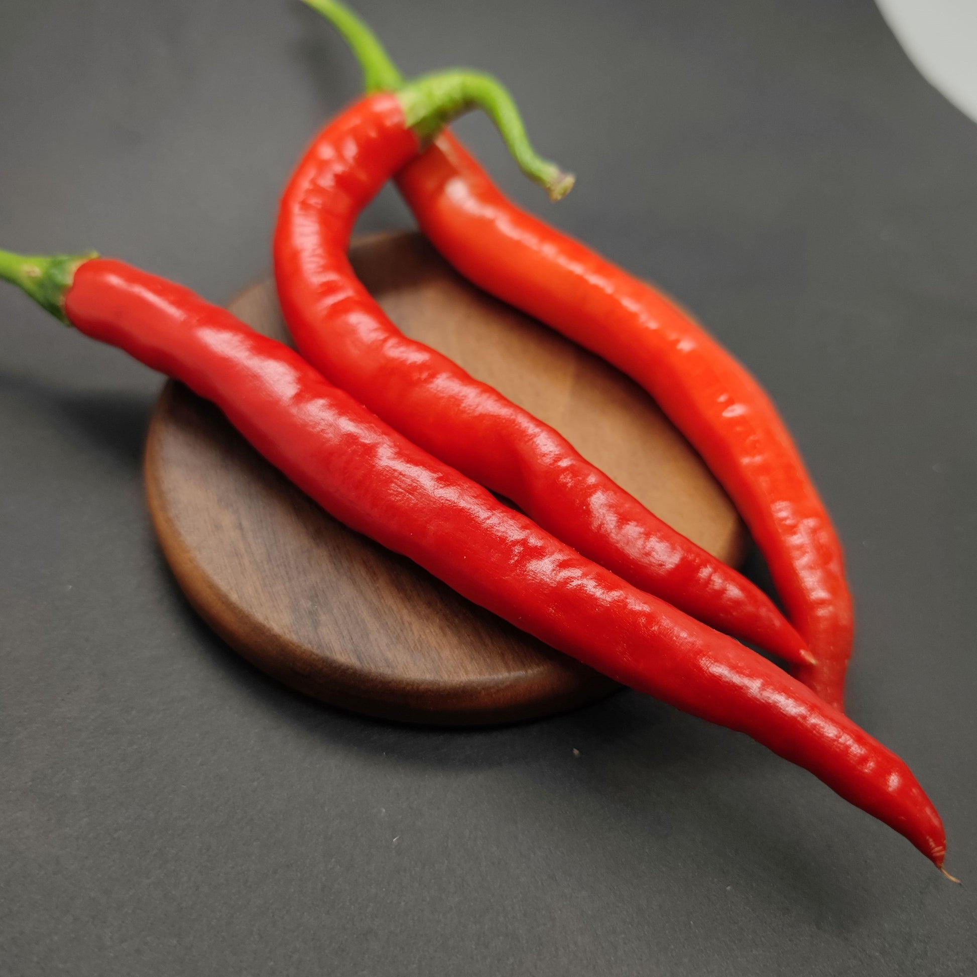 Three vibrant Hangjiao 10 Afterglow (HJ10) peppers from PepperMerchant.net are placed on a round, wooden tray. The chilies curve naturally and have green stems. The background is dark and contrasts with the bright red color of the peppers, highlighting their glossy texture and showcasing space innovation in agriculture.