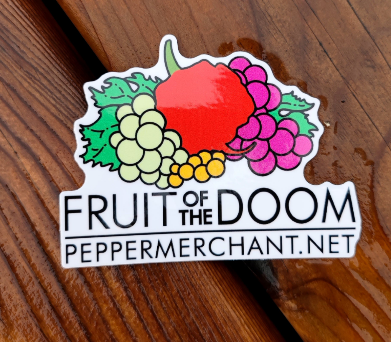 The Fruit of The Doom Sticker by PepperMerchant.net features a colorful parody design with grapes, a red pepper, and leaves above the text “FRUIT OF THE DOOM” and “peppermerchant.net,” shown on a wooden surface.