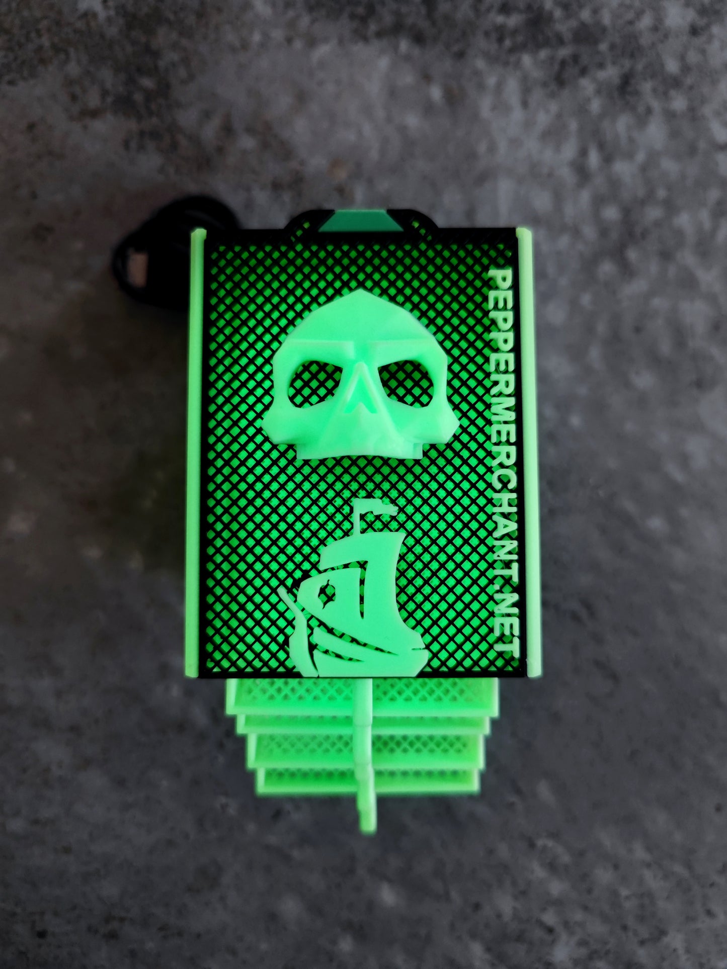 A green pepper spray canister featuring a skull and ship graphic, “PepperMerchant.net” text, and a tribute to the Seed Dryer - Limited Glowing Edition, displayed on a textured gray background.