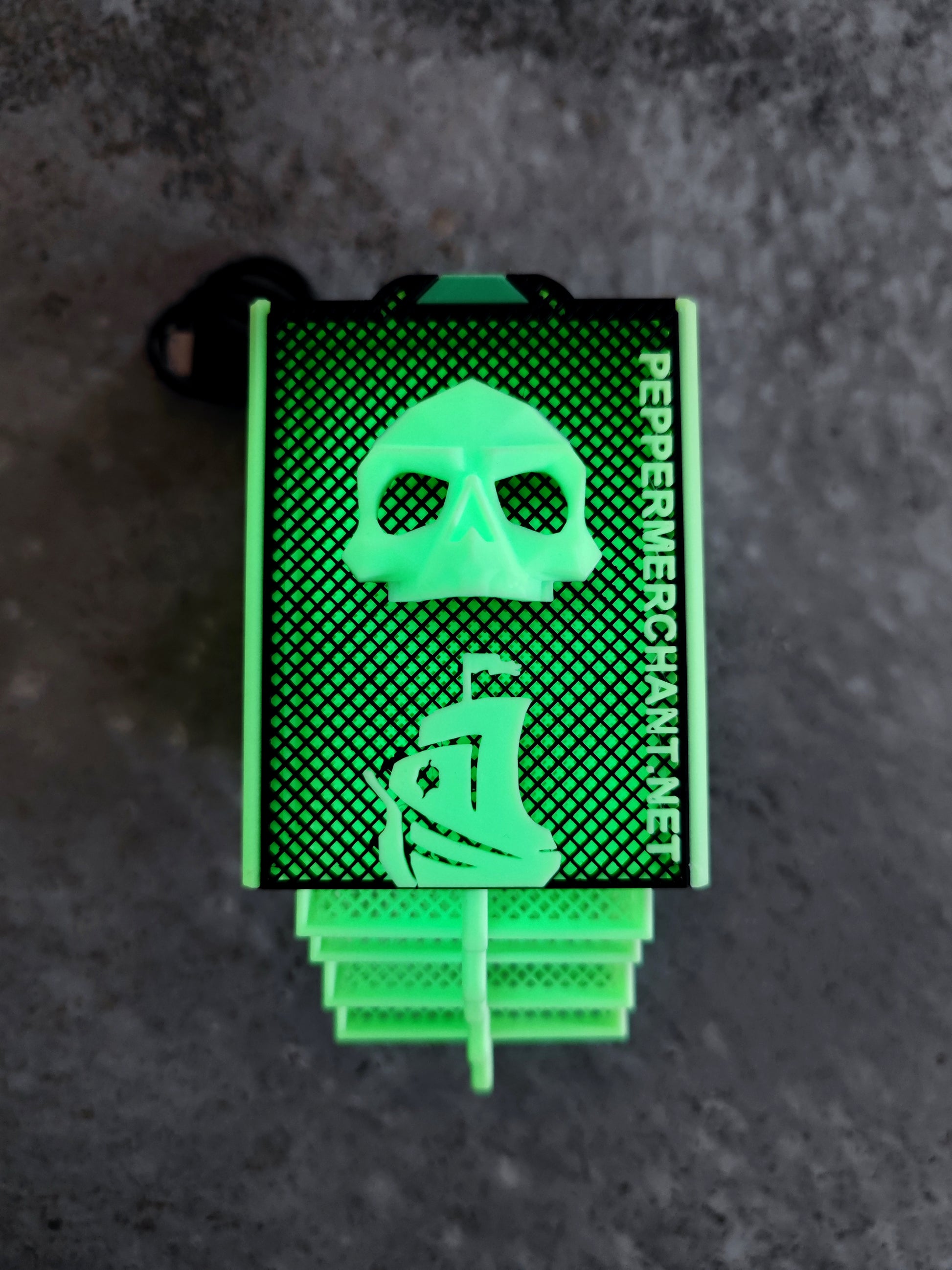A green pepper spray canister featuring a skull and ship graphic, “PepperMerchant.net” text, and a tribute to the Seed Dryer - Limited Glowing Edition, displayed on a textured gray background.