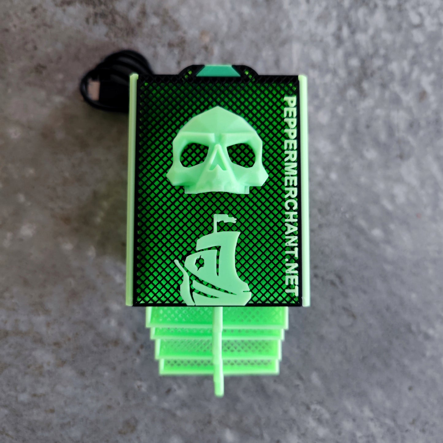 The Seed Dryer - Limited Glowing Edition by PepperMerchant.net features a green and black rectangular design with skull and pirate ship art, "PEPPERMERCHANT.NET" branding, and sits on a gray textured surface—a unique blend of style and seed-saving tech.