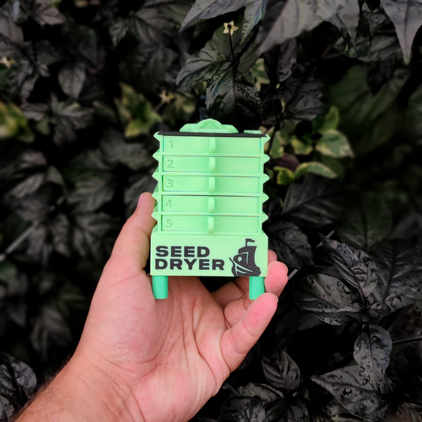 A hand holds a small, stackable "Seed Dryer - Limited Glowing Edition" by PepperMerchant.net with trays numbered 1–5. The green dryer stands out against a backdrop of dark leafy plants.