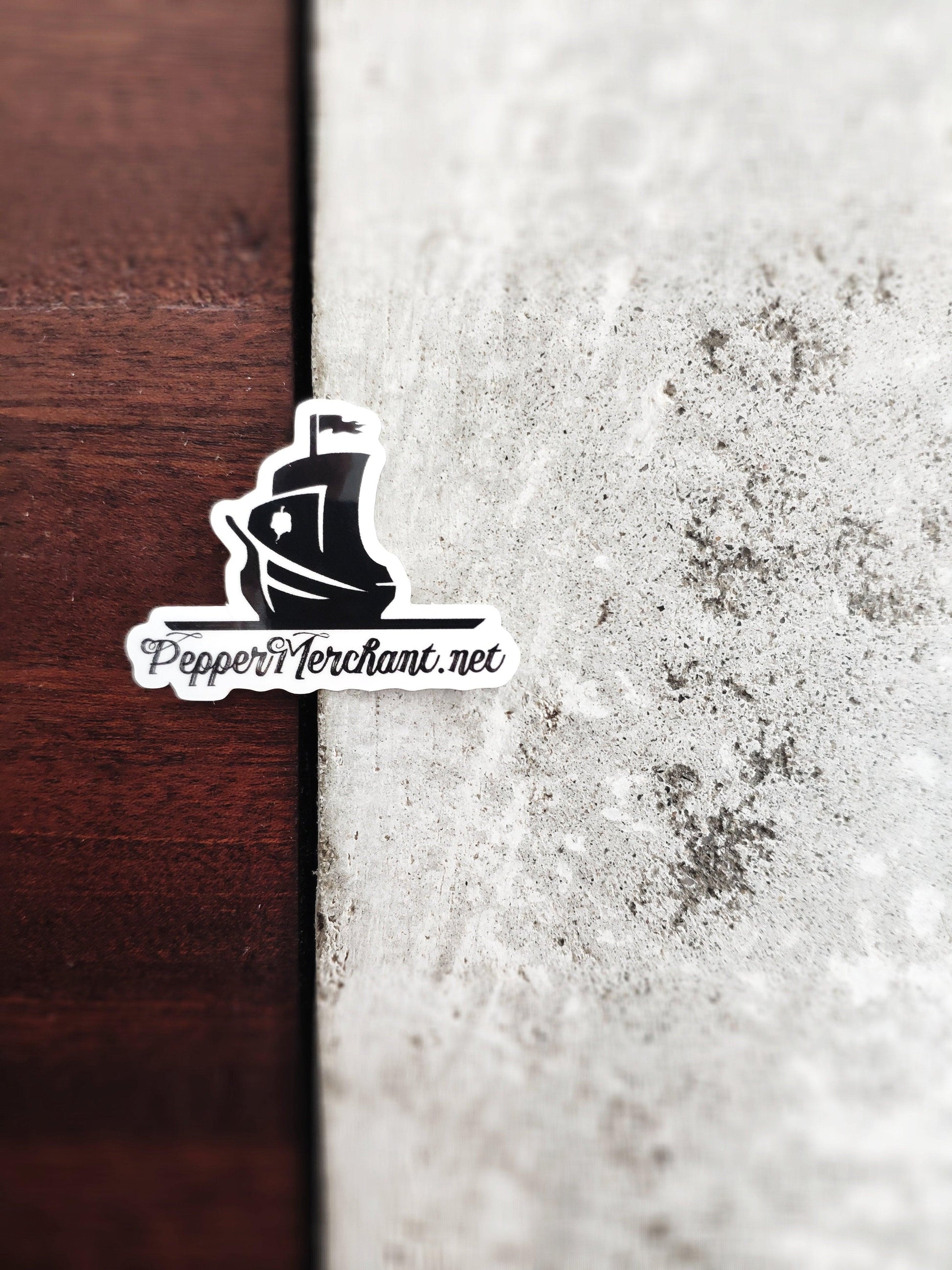 A PepperMerchant.net Sticker, showcasing a black ship silhouette with a prominent mast and birdlike emblem alongside the text "PepperMerchant.net," is affixed where wooden and concrete textures converge.