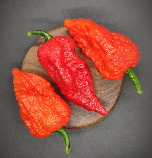 Three vibrant red "Yellow Fever - Red (Death Cheeto)" peppers from PepperMerchant.net, featuring wrinkled, textured skin, are arranged on a small round wooden board with a dark background. The peppers look fresh, their green stems still attached, vividly reminding one of nature's own Death Cheeto.