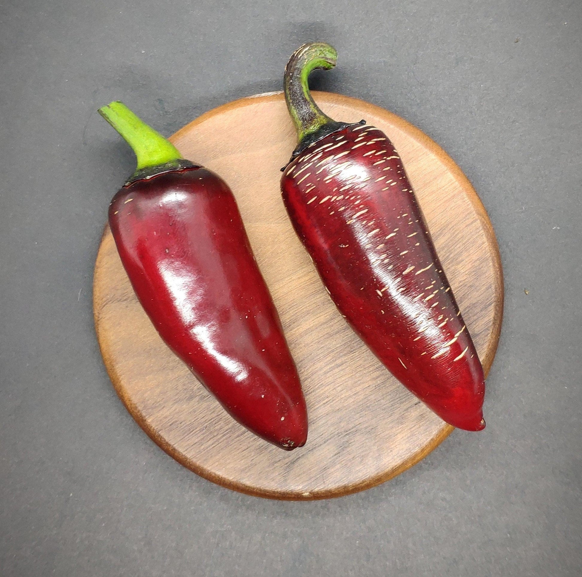 Two vibrant red Ruby Jalapeno peppers from PepperMerchant.net rest on a round wooden coaster against a dark background. The pepper on the right showcases brown streaks that indicate its ripeness. With green stems and textured wood grain from the coaster adding depth to the image, these unique pepper seeds are perfect for those seeking something special at an affordable price.