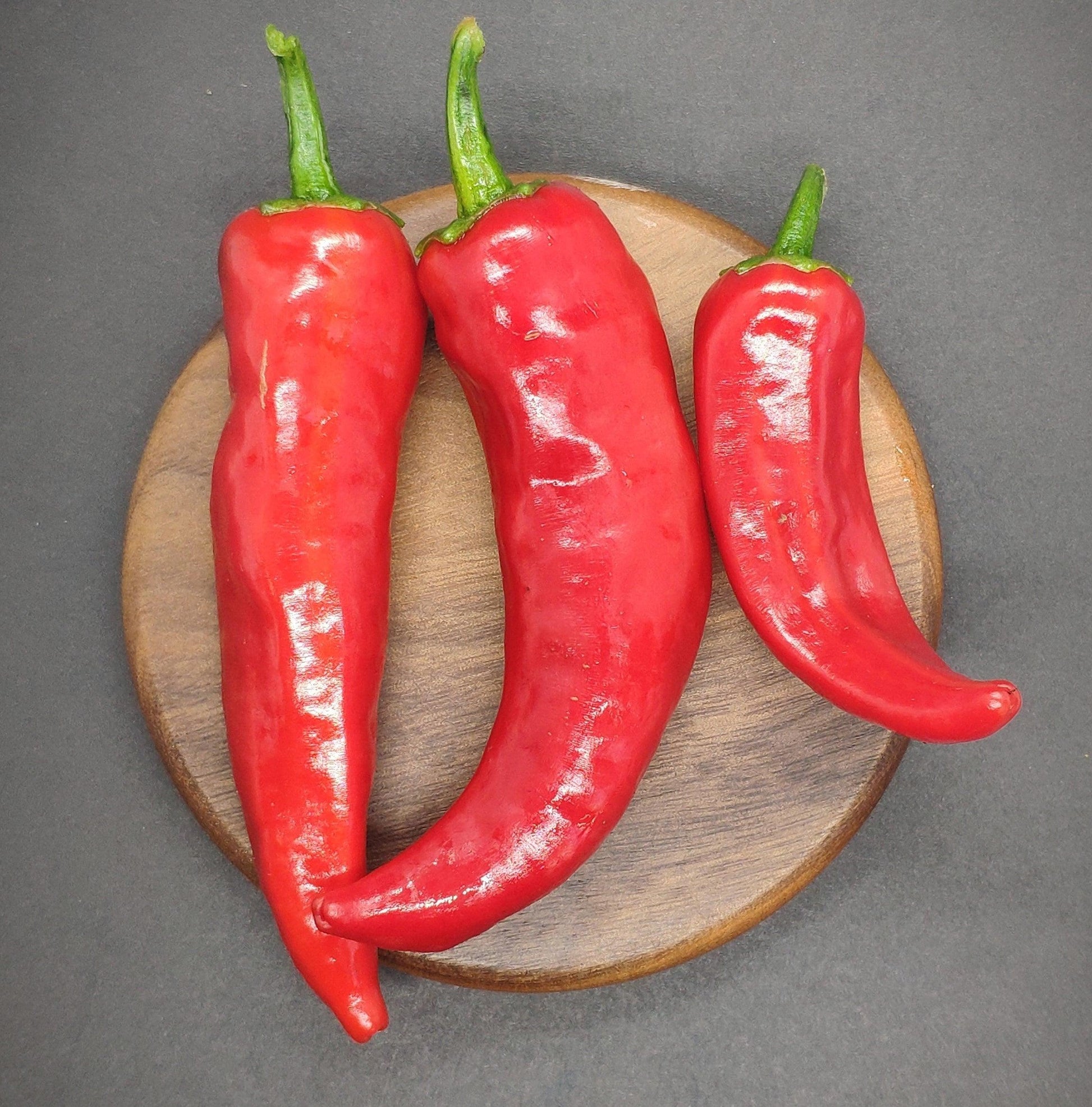 Three glossy red Fish Peppers, celebrated for their culinary versatility, are showcased on a round wooden board against an elegant dark background. The peppers vary in size; two larger ones positioned on the left and a smaller one on the right. Each pepper features a green stem and boasts a vibrant red hue, indicative of their moderate heat level. These exquisite peppers are brought to you by PepperMerchant.net.