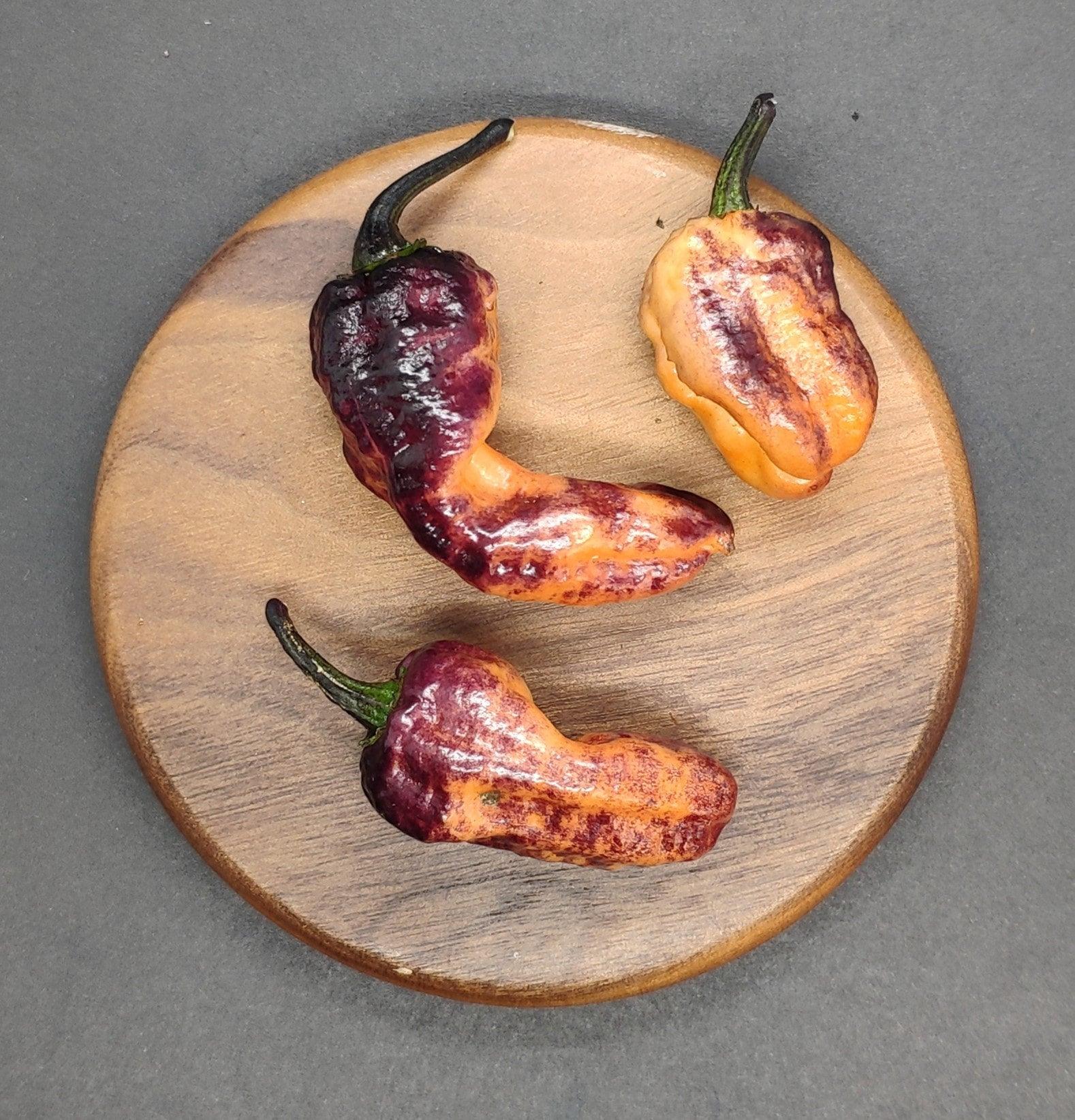 A wooden plate showcasing three medium-sized chili peppers, predominantly in purple and orange hues with a slight charred texture, features the PepperMerchant.net exclusive Black Ruby Pink peppers set against a gray background.