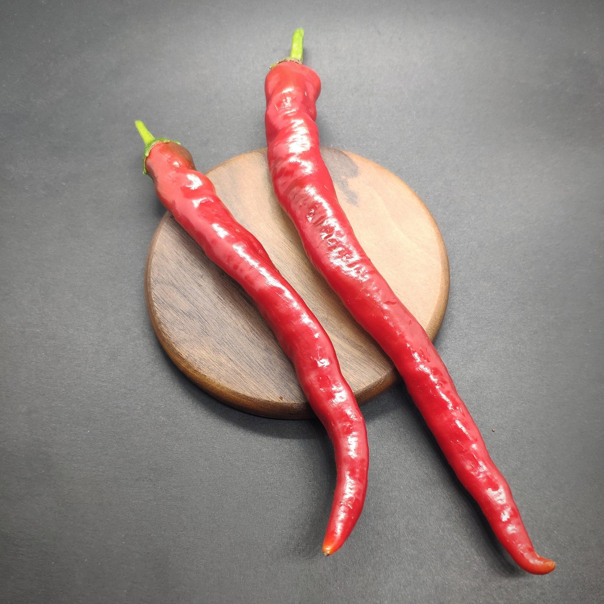 Two long, red Hangjiao 7 Supernova peppers from PepperMerchant.net, with green stems, are placed side by side on a round wooden board against a dark background. The Hangjiao 7 Supernova pepper seeds within promise intense heat. The chilies are slightly curved and have a glossy surface, radiating an almost celestial beauty.