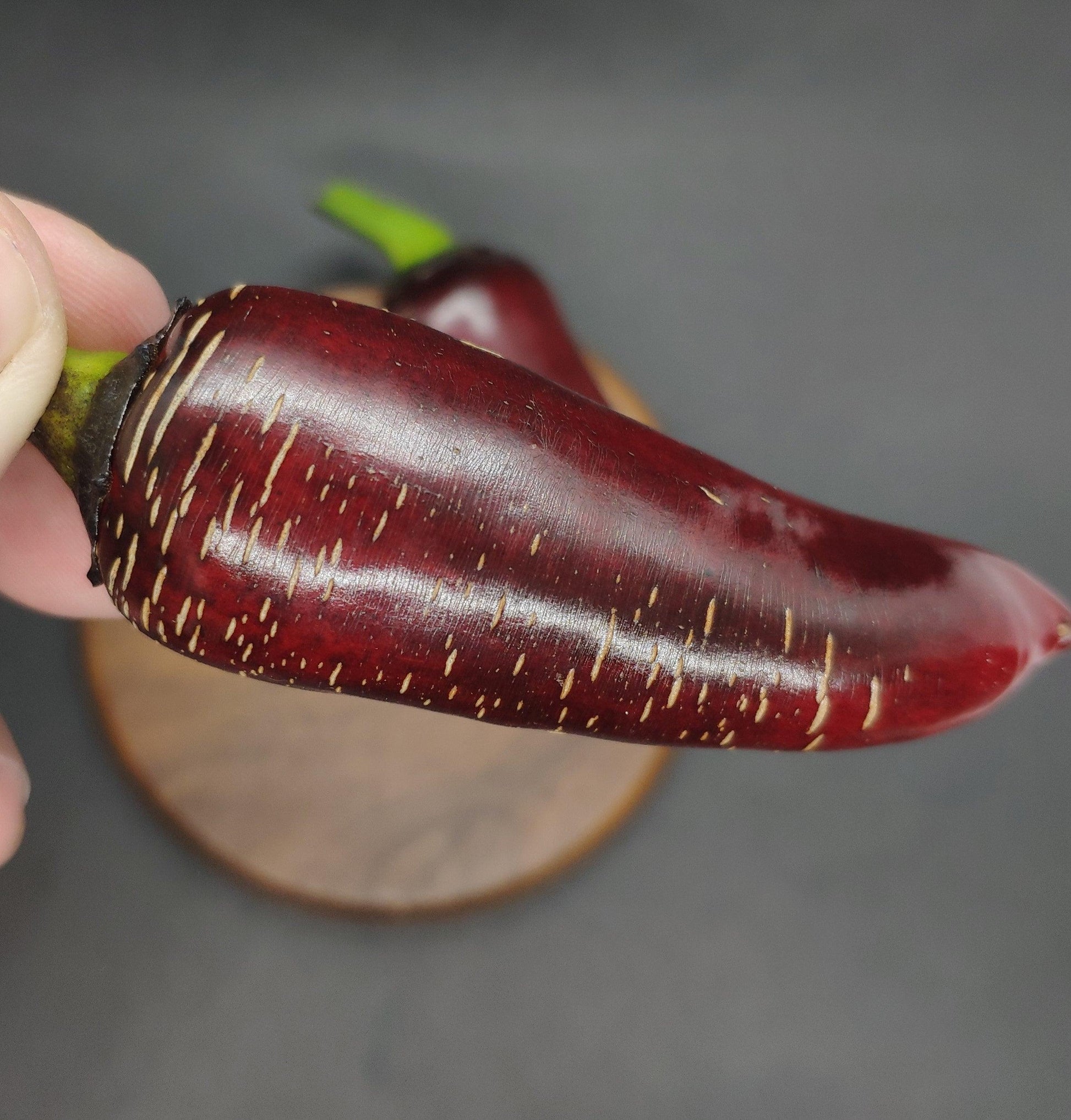 A close-up image showcases a shiny, deep red Ruby Jalapeno from PepperMerchant.net, delicately held between two fingers. The chili pepper features distinctive light-colored streaks running along its surface. In the background, another Ruby Jalapeno rests on a wooden cutting board. These Ruby Jalapenos come with unique seeds perfect for your garden.