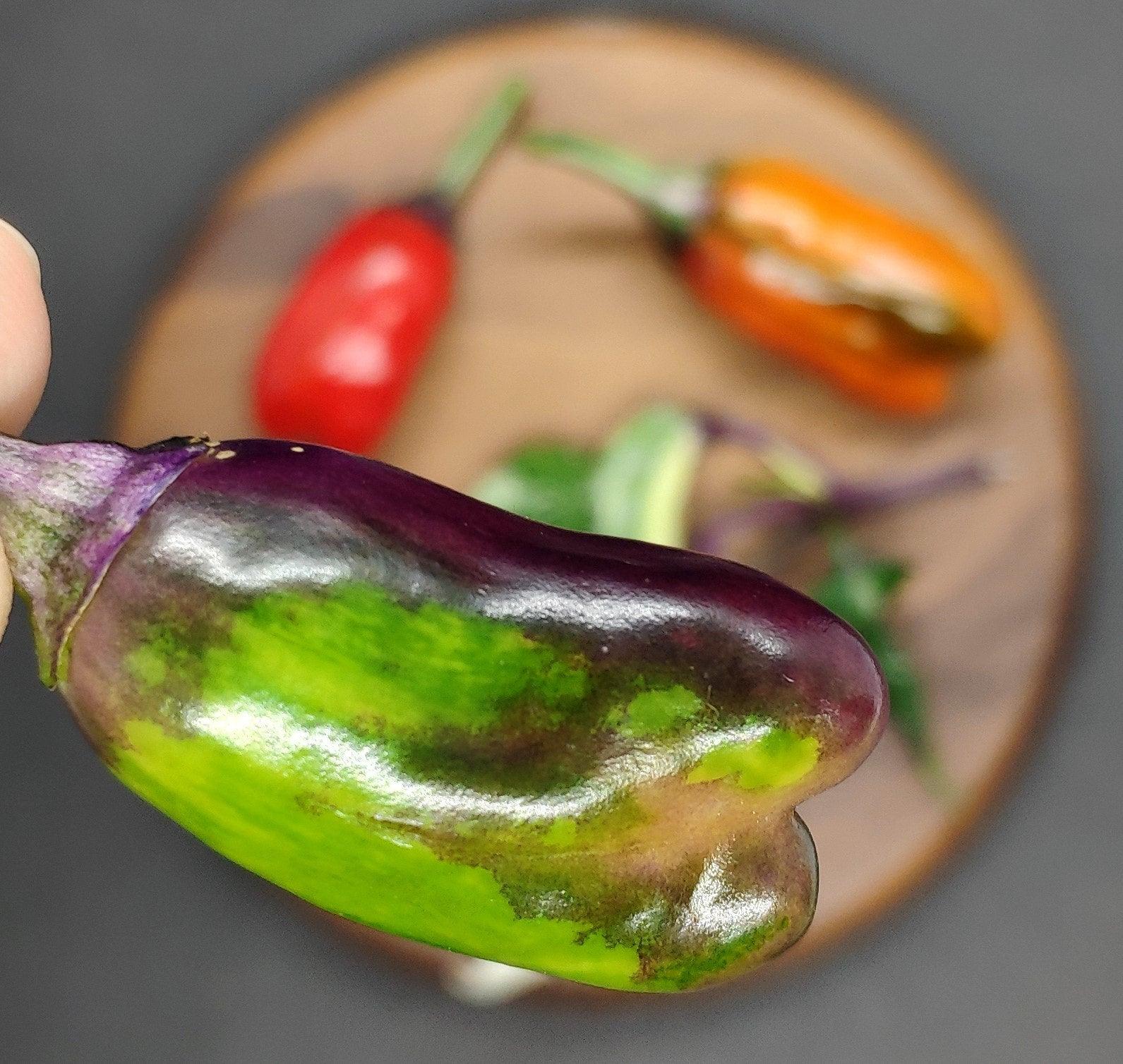 Close-up of a hand holding a uniquely shaped, partially purple and green RH Lilac pepper from PepperMerchant.net, resembling the ornamental pods found in their gardens. In the blurred background, there's a brown plate with various colorful RH Lilac peppers and greens on it.