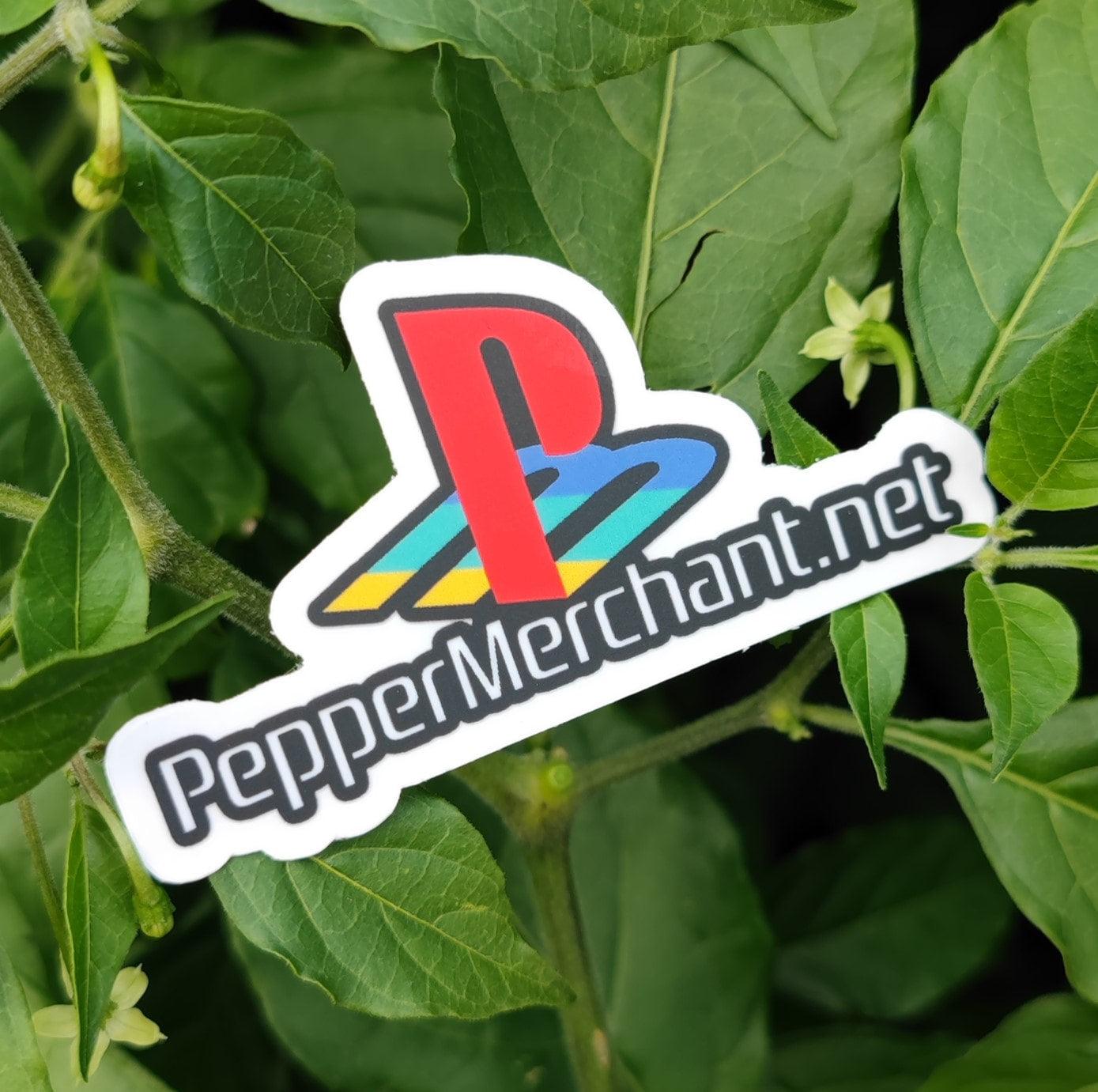 A PepperStation Sticker from PepperMerchant.net, featuring a PlayStation-inspired logo with gradients of red, blue, and yellow, is placed among lush green leaves. This colorful parody design is perfect for true gamers.