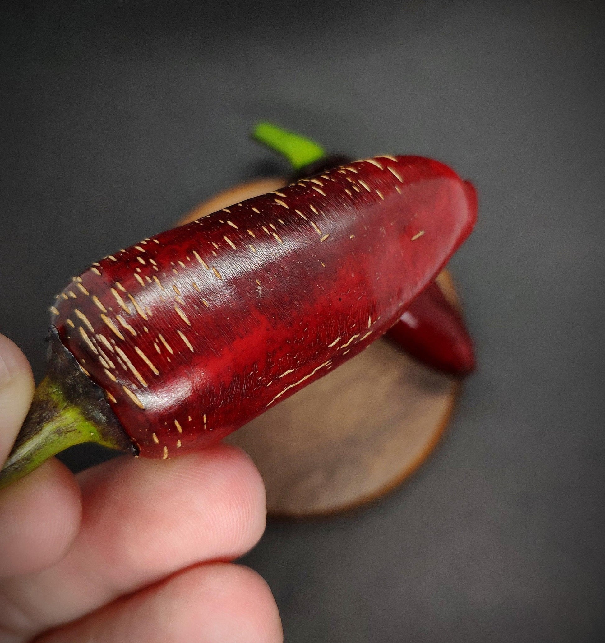 A close-up image showcases a person holding a dark red Ruby Jalapeno from PepperMerchant.net, with small streaks of lighter color. The blurred background accentuates the vibrant color and texture of the chili pepper, while another Ruby Jalapeno and a wooden surface are also visible in the background.