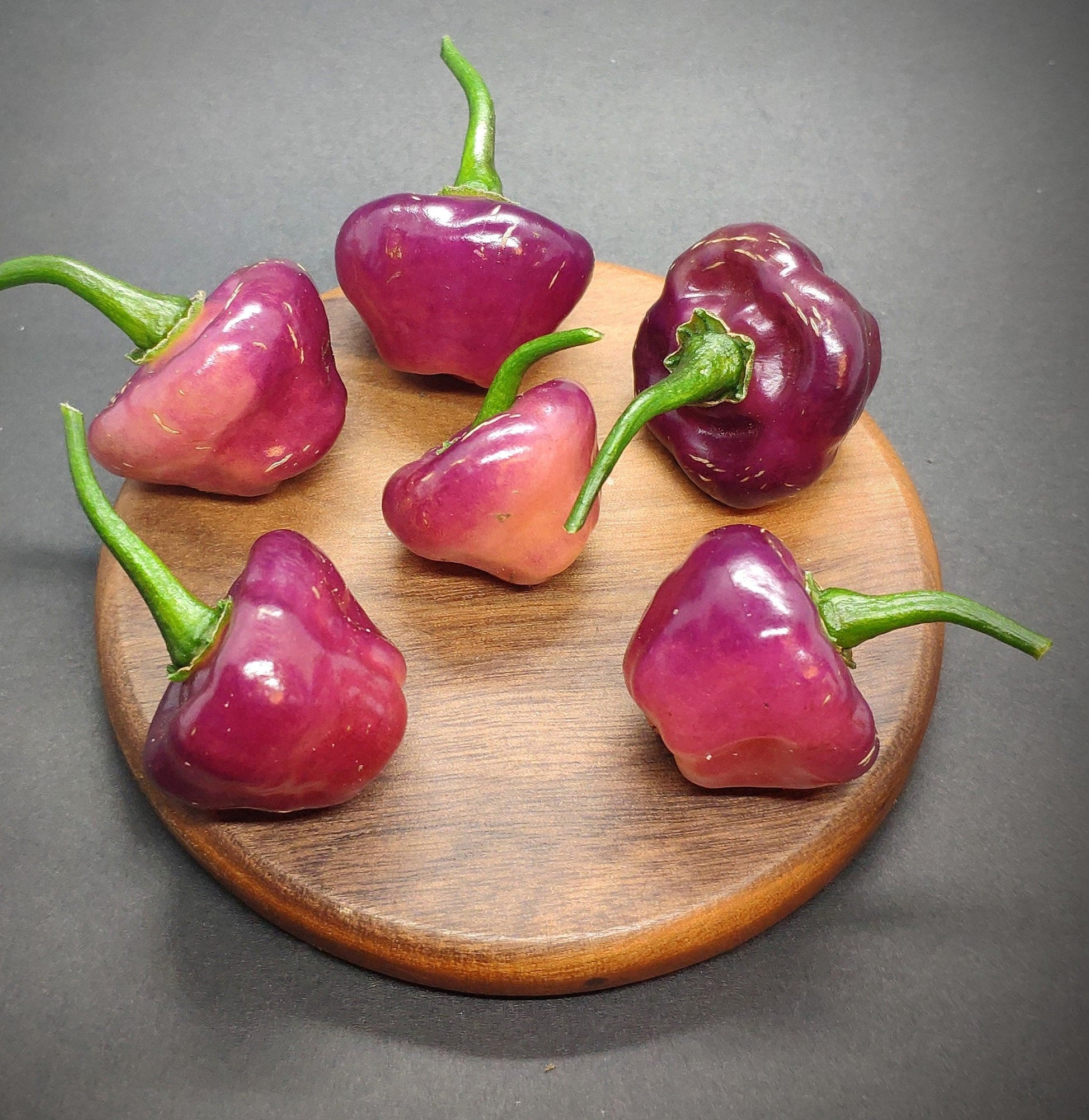 Five Cheiro Roxa chili peppers with green stems are arranged on a round wooden board, set against a dark background. The glossy peppers from PepperMerchant.net, known for their mild flavor, vary in shape and add a visually rich contrast to the wood. Hailing from Brazil, they introduce an exotic twist to the scene.