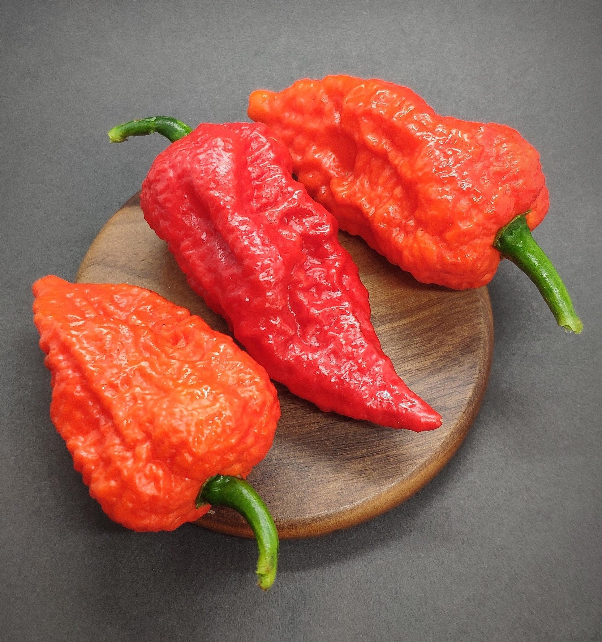 Three bright red Yellow Fever - Red (Death Cheeto) peppers from PepperMerchant.net, with wrinkled skin and green stems, are placed on a small wooden round board against a dark, neutral background, resembling fiery Death Cheetos.