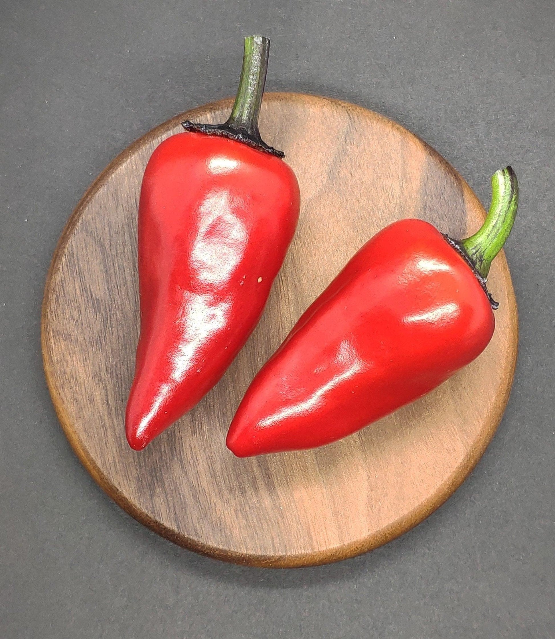 Two fresh, shiny Count Dracula 🧛 peppers with green stems rest on a round wooden plate against a dark background, showcasing the productive plant's early harvest from PepperMerchant.net Count Dracula 🧛 Pepper Seeds.