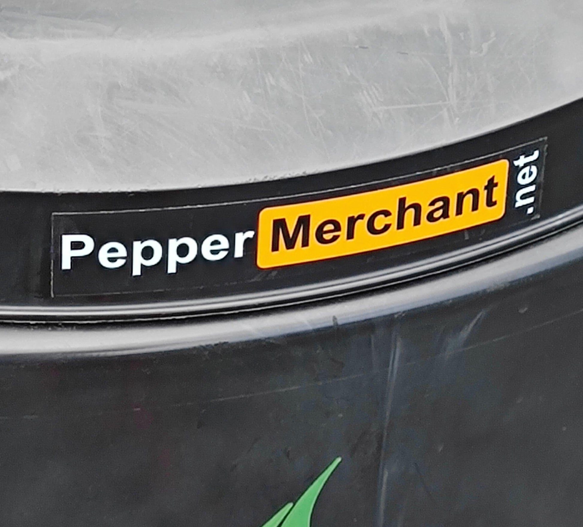 A close-up image of a black container adorned with a "Pepper Hub Sticker" from PepperMerchant.net. The background seems to be a metallic surface. The sticker displays the website name in white font, with "Merchant" highlighted in yellow, providing it with a parody-like appearance.