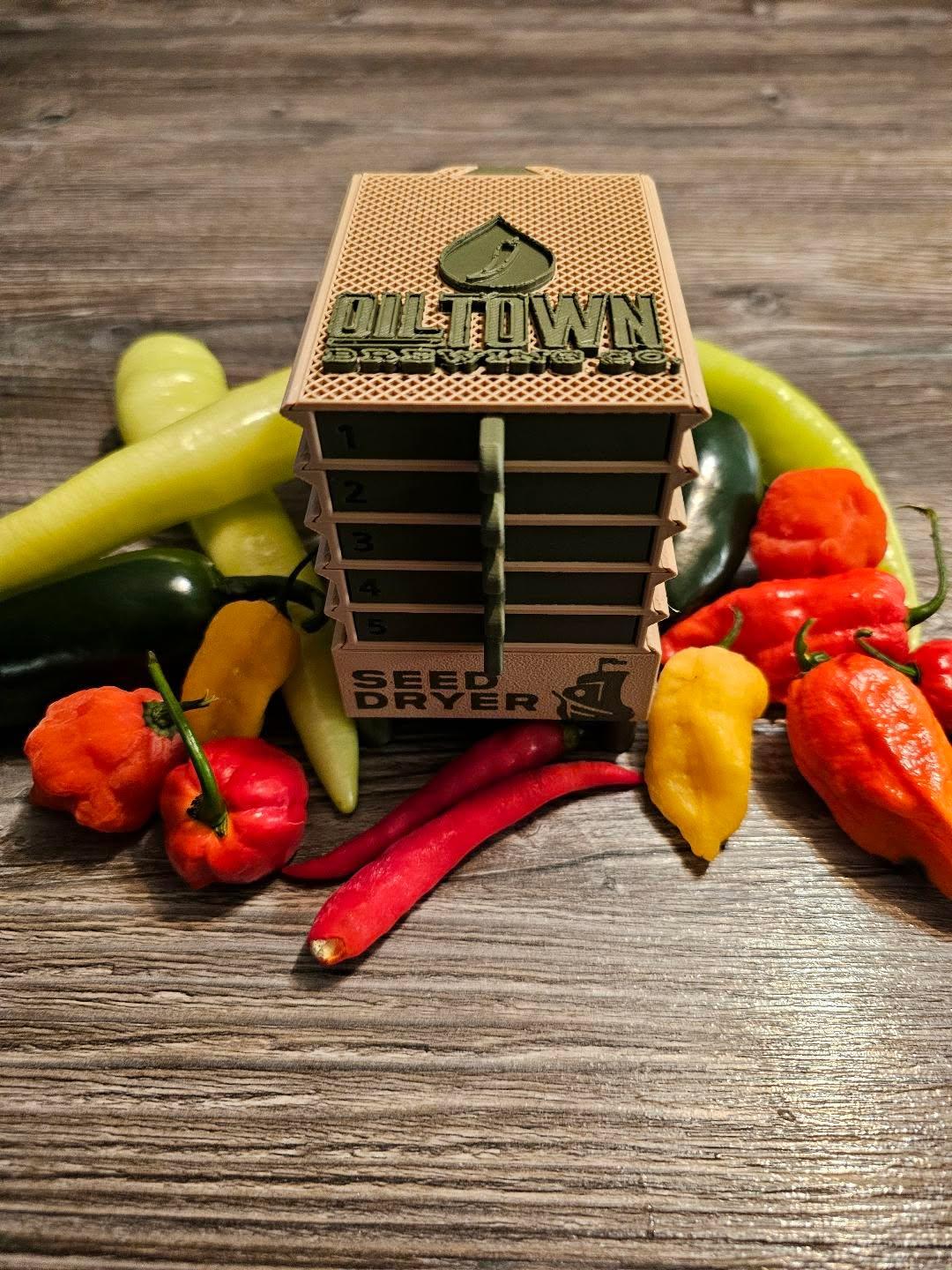 A compact, tan Seed Dryer - Custom Design by PepperMerchant.net sits on wood, surrounded by colorful peppers. This USB-powered dryer features innovative seed-saving tech in addition to traditional drying capabilities.