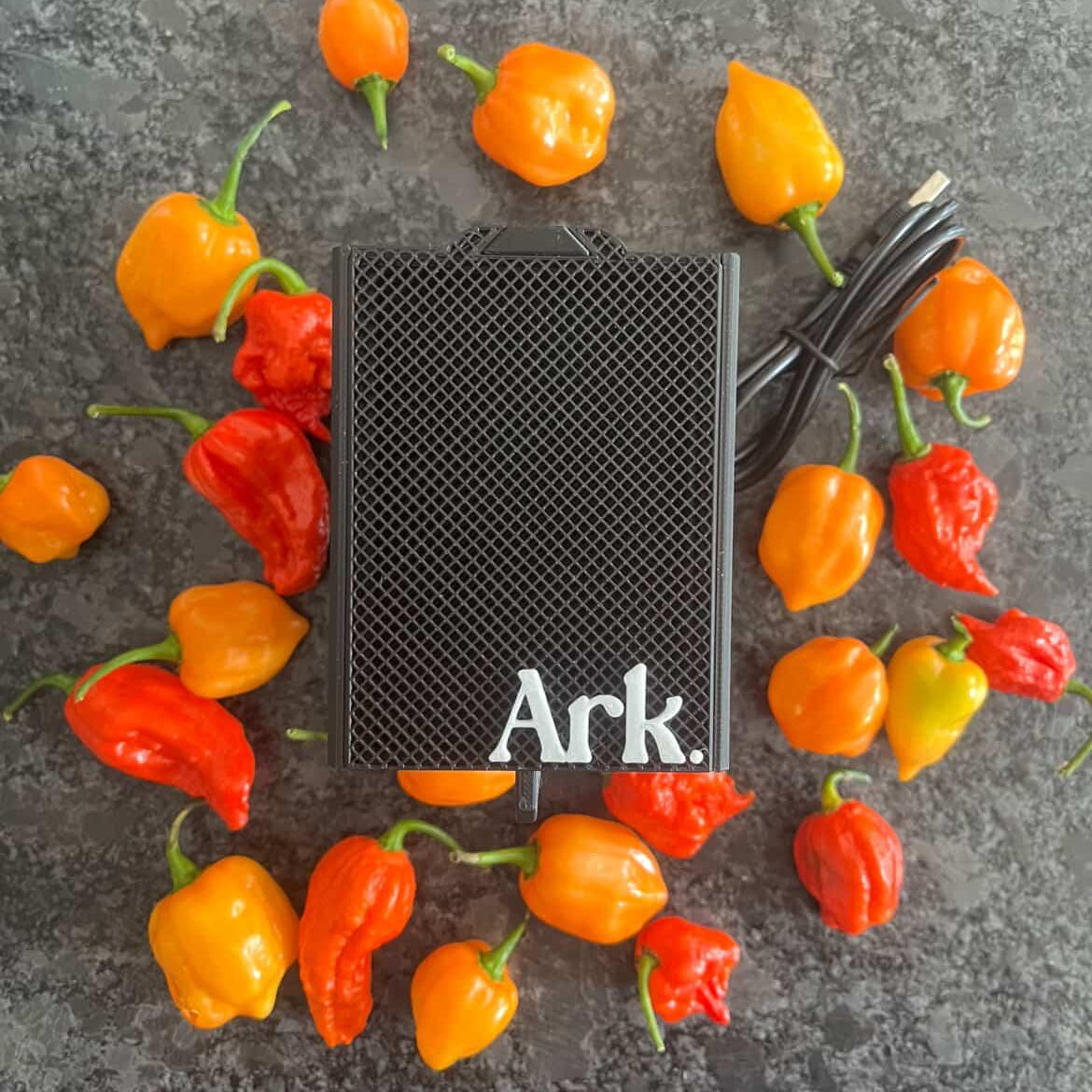 The Seed Dryer - Custom Design by PepperMerchant.net, a sleek black USB-powered device, sits on a dark textured surface among orange and red hot peppers, with its black power cord alongside.